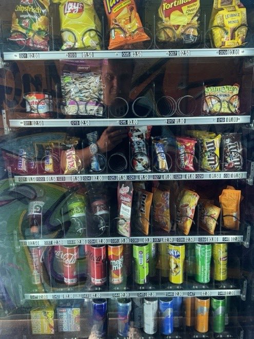 Blick durch die Scheibe auf ein volles Automatenregal mit Chips, Süßem und Getränken