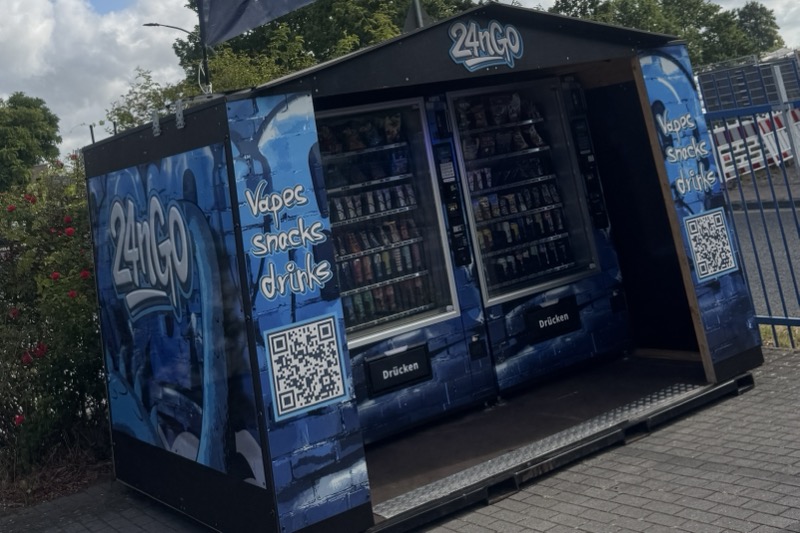 Blauer 24nGo-Ministore mit geöffneter Front, dahinter zwei Automaten mit Getränken und Snacks